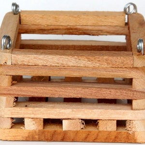 May include: A square wooden crate constructed from horizontal slats with a natural light brown finish. The structure features four silver metal eye hooks attached to the top corners, allowing for suspension. The design is open and slatted.