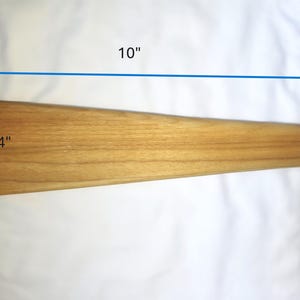 May include: A tapered wooden block with a smooth, light brown grain. The object is marked with blue dimension arrows indicating a length of 10 inches and a width of 2 3/4 inches at the widest end, showing a clear, rectangular profile.