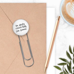 May include: A white button with the text "I'm silently correcting your grammar." attached to a silver paper clip. The button is on a brown envelope.
