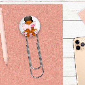 May include: A pink notebook with a silver bookmark featuring a woman reading a book with a pink heart on the cover. The bookmark is attached to the notebook with a silver clip.