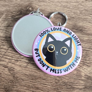 May include: A pink and purple round button with a black cat illustration. The button has the text "100% Love and Light But Don't Mess With Me".