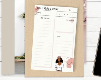 May include: A cream-colored notepad with the text "GET THINGS DONE" and a to-do list layout. The notepad features a minimalist illustration of a person holding a coffee cup. The notepad is on top of a beige book and next to a smartphone.