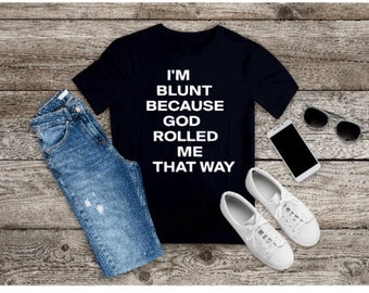 May include: A black short-sleeved t-shirt with white text that reads I'M BLUNT BECAUSE GOD ROLLED ME THAT WAY, paired with blue denim jeans, white sneakers, a smartphone, and dark-tinted sunglasses arranged on a rustic wooden surface.