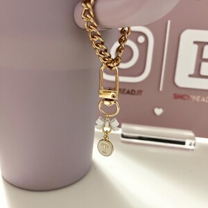 May include: A gold chain with a gold clasp and a white charm with a gold letter "T" and two white bows. The chain is attached to a purple mug.