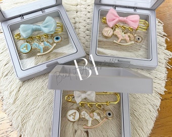 Può includere: Tre scatole bianche con coperchi trasparenti, ciascuna contenente una spilla per neonati dorata con un fiocco blu, rosa o bianco, un ciondolo a forma di cavallo a dondolo, un ciondolo con una lettera e un ciondolo con un occhio malocchio.