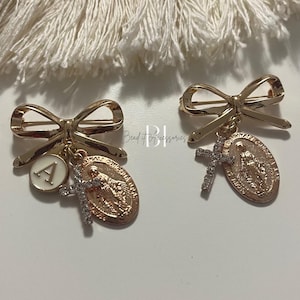 May include: Two gold-toned brooches shaped like bows with charms. The charms include a cross with clear rhinestones, a round charm with a religious image, and a round charm with the letter "A".