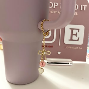 May include: A gold chain with a pink and white bow charm, a pink circle charm with the letter "T" and a white heart charm. The chain is attached to a purple tumbler.