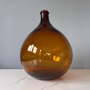 May include: A large, round, amber-colored glass carboy or demijohn bottle with a narrow neck and a thick, rounded rim. The translucent glass features a smooth, reflective surface that catches light, showcasing a deep, warm brown hue throughout.