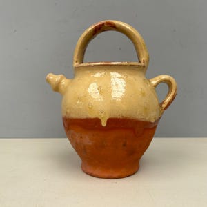 Puede incluir: Una jarra de cerámica hecha a mano con asa y boquilla. La jarra es de un color cálido y terroso, con una parte superior de color amarillo claro y una base marrón rojizo. El asa es arqueada y la boquilla es pequeña y curvada.