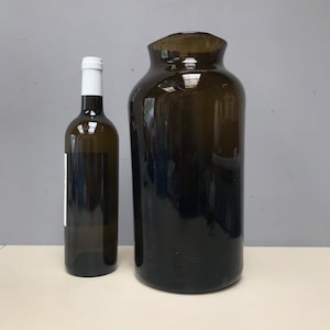 Puede incluir: Un jarrón de vidrio marrón oscuro con una boca ancha y una base estrecha, junto a una botella de vidrio transparente con una etiqueta blanca.