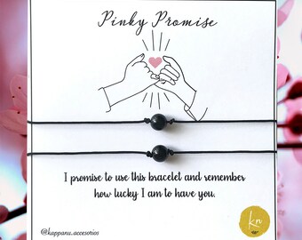 Puede incluir: Dos pulseras de cuerda negra con cuentas de ónix negro. Las pulseras se muestran en una tarjeta blanca con el texto "Pinky Promise" y un dibujo de dos manos haciendo una promesa con el meñique. La tarjeta también incluye el texto "Prometo usar esta pulsera y recordar lo afortunado que soy de tenerte." y el texto "@kappanu.accesorios".
