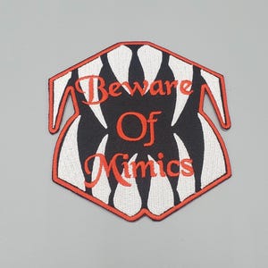 May include: Embroidered patch with a stylized design featuring a black and white pattern resembling teeth, outlined in red. The text "Beware Of Mimics" is in red, with a playful, slightly gothic font.