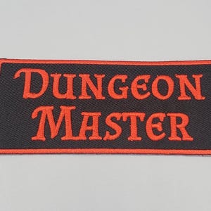 May include: A rectangular patch with the words "DUNGEON MASTER" embroidered in orange thread on a black background. The patch has an orange border and is likely for clothing or accessories.