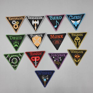 May include: A collection of triangular embroidered patches featuring various fantasy class names like "Artificer," "Barbarian," and "Wizard." Each patch has a unique color scheme and symbol representing the class. The patches are on a light gray surface.
