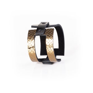 Puede incluir: Un brazalete de cuero dorado y negro con un diseño texturizado. El brazalete está hecho de dos capas de cuero, una dorada y una negra. La capa dorada tiene un diseño texturizado que se asemeja a escamas. La capa negra es lisa y tiene un cierre de hebilla.