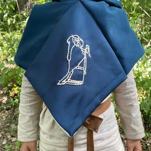 May include: A navy blue hooded cloak featuring a white embroidered design. The cloak is worn over a light-coloured shirt and a brown leather belt. The background is a natural outdoor setting with green foliage.