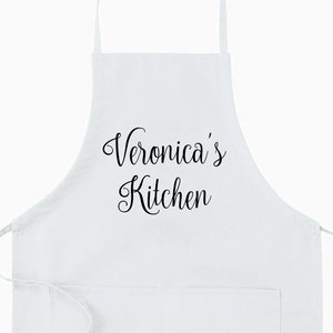 以下が含まれることがあります： 大きなポケットと調節可能なストラップが付いた白いキッチンエプロン。エプロンには、「Veronica's Kitchen」という文字が前面に印刷されています。