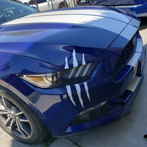 Op de afbeelding: Een blauwe Ford Mustang met witte race strepen en een witte klauwmarkering op de voorvleugel.