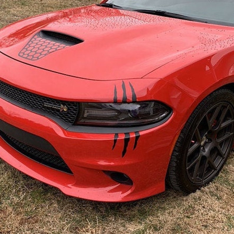 Op de afbeelding: Een rode Dodge Charger met zwarte klauwen decals op de voorbumper. De auto heeft een zwart honingraat grille en zwarte wielen.
