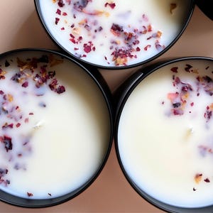 May include: Three round, black candles with white wax and dried rose petals. The candles are in black tins and have a single wick. The petals are red and tan, scattered across the surface of the wax.