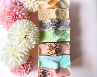 May include: A stack of handmade soaps in various colors and textures, including light green, pink, and blue. Some soaps are topped with decorative elements like dried flowers and seeds. The soaps are arranged vertically next to white and pink pom-pom flowers.
