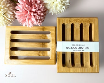 May include: Two rectangular bamboo soap dishes. The larger dish has the text "ECO-FRIENDLY BAMBOO SOAP DISH SoulSoapUsa" on a white label. Both dishes have a slatted design.