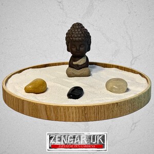 May include: A miniature Zen garden featuring a small Buddha statue, three polished stones, and white sand in a round bamboo tray. The Buddha is brown, and the stones are yellow, black, and clear. The tray is approximately 25 cm in diameter.
