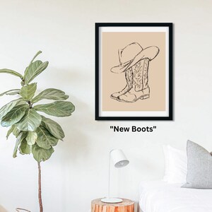 May include: A black framed print with a beige background featuring a line drawing of a cowboy hat and boots. The text "New Boots" is below the image.