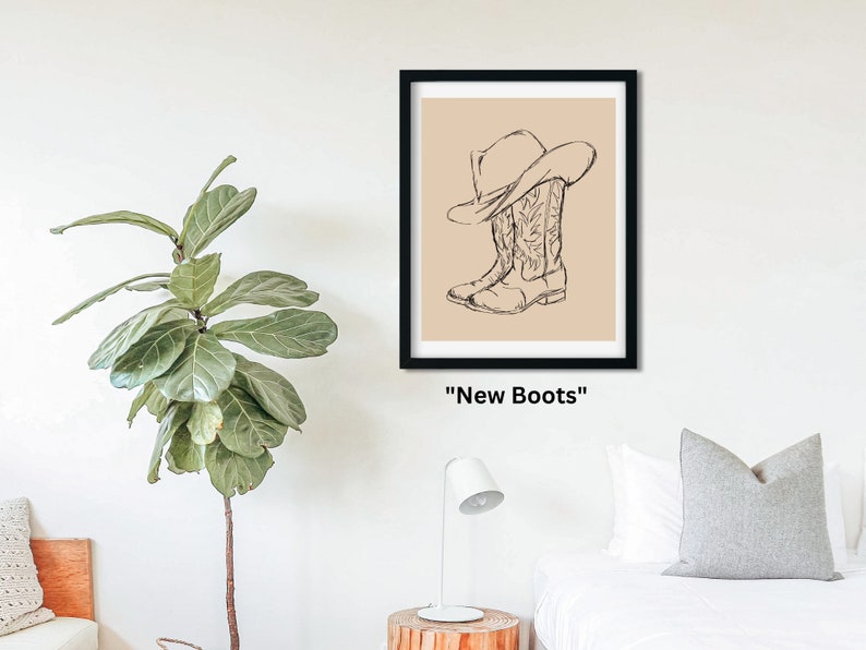 May include: A black framed print with a beige background featuring a line drawing of a cowboy hat and boots. The text "New Boots" is below the image.