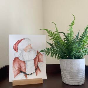 May include: Watercolor painting of Santa Claus wearing a red suit and white beard, framed and displayed on a wooden easel. A potted fern plant is on the right side of the image.