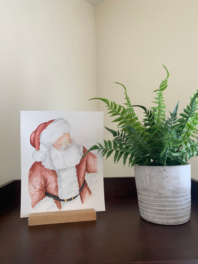 May include: Watercolor painting of Santa Claus wearing a red suit and white beard, framed and displayed on a wooden easel. A potted fern plant is on the right side of the image.