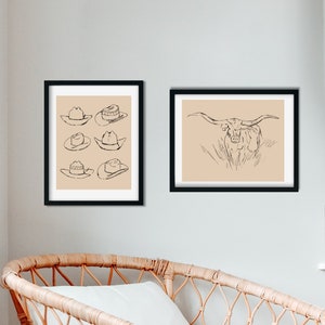 May include: Two black framed prints on a white wall. The print on the left features a line drawing of six cowboy hats. The print on the right features a line drawing of a longhorn steer.