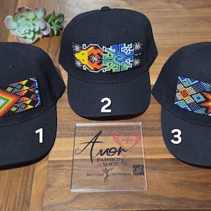 Puede incluir: Tres gorras de béisbol negras con parches rectangulares de cuentas con patrones geométricos coloridos. Cada gorra está numerada uno, dos o tres. Un letrero cuadrado transparente muestra el texto Amor Fashion Shop Boutique Artisanal y un código QR.