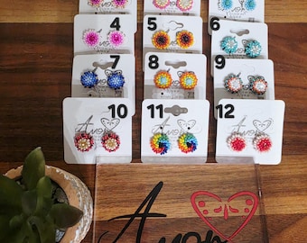 May include: Twelve pairs of round, multi-colored beaded stud earrings displayed on white cards numbered one through twelve. The cards feature the text Amor Fashion Shop Boutique Artesanal and a heart graphic. Each pair features intricate beadwork.