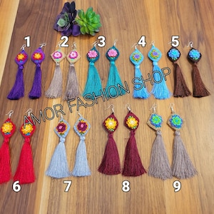 May include: A collection of nine pairs of handmade crochet tassel earrings in various colors. Each earring features a floral design at the top and a long tassel. The earrings are displayed on a wooden surface, with the text "AMOR FASHION SHOP" visible.