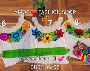 May include: Three white embroidered tops with colorful floral designs. The tops have a white lace trim and are labeled with the numbers 6, 7, and 8. The text "AMOR FASHION SHOP" and "Embroidered" are visible. A green sash and a colorful embroidered pouch are also included. The text "BUST 36/38" is visible.