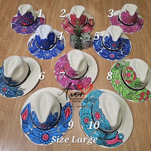 Peut inclure: Dix chapeaux de paille peints à la main, chacun avec un motif floral unique de différentes couleurs. Les chapeaux ont une couronne et un bord blancs, avec des motifs peints sur le bord. Le texte "Size Large" est visible.