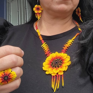May include: A beaded jewelry set featuring a sunflower-shaped pendant necklace, matching flower-shaped drop earrings, and a flower-shaped ring. The pieces are crafted with small yellow, orange, red, and black beads in a vibrant floral pattern.