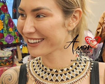 May include: A wide, circular beaded collar necklace featuring a geometric diamond pattern in black, white, and metallic gold beads. The lower edge is finished with long, dangling fringe strands of black and gold beads with small white bead accents.