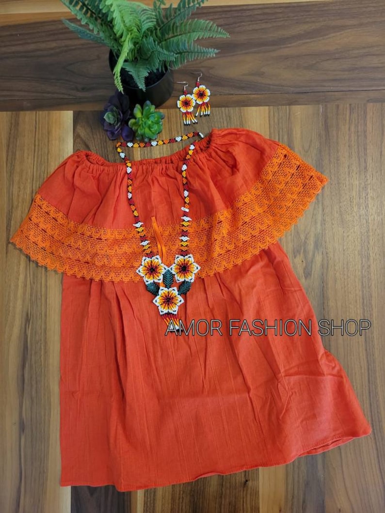May include: An orange off-the-shoulder blouse featuring a wide lace trim along the neckline, paired with a matching beaded necklace and earrings set. The jewelry displays a floral pattern in white, yellow, orange, and black. Text reads AMOR FASHION SHOP.