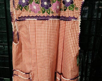 May include: An orange and white gingham apron featuring a scoop neckline and two front pockets. The apron is decorated with embroidered purple and pink flowers with green leaves and finished with a purple crocheted trim along the edges.