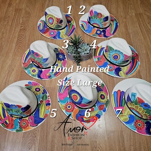 Puede incluir: Sombreros de sol blancos, pintados a mano, talla grande, con coloridos diseños florales y de pájaros. Cada sombrero presenta una corona blanca y un ala ancha adornada con patrones vibrantes en tonos de azul, amarillo, rosa y verde. Los sombreros se exhiben sobre una superficie de madera.