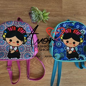 Puede incluir: Dos mochilas pequeñas con un personaje de dibujos animados de pelo oscuro y diadema floral. Una mochila es rosa con estampado de mandalas y la otra es azul con estampado de hojas. Ambas tienen correas ajustables y un gráfico central.