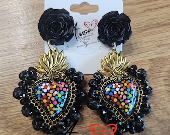 May include: A pair of earrings featuring black rose studs attached to heart-shaped pendants. The pendants have a gold-tone metal frame, a central black heart with colorful polka dots, and a border of black beads. The text Amor Fashion Shop is visible.