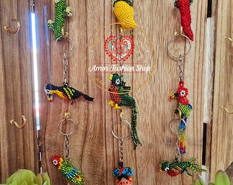 May include: A collection of eight beaded keychains hanging on a wooden display. The designs include a green cactus, a yellow mango, a red chili pepper, and five colorful birds, plus one figure wearing a dress, all crafted with small beads.