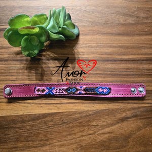 Könnte beinhalten: Ein pinkfarbenes Lederarmband mit einem Perlendesign in Blau, Braun und Weiß. Das Armband hat silberfarbene Druckknöpfe. Die Worte "Amor Fashion Shop" sind im Bild.