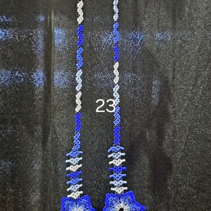 May include: A long beaded necklace featuring a floral pendant with three blue, white, and green flowers. The necklace has two long strands of blue and white beads, with two smaller matching flower accents at the top. The number 23 is visible.