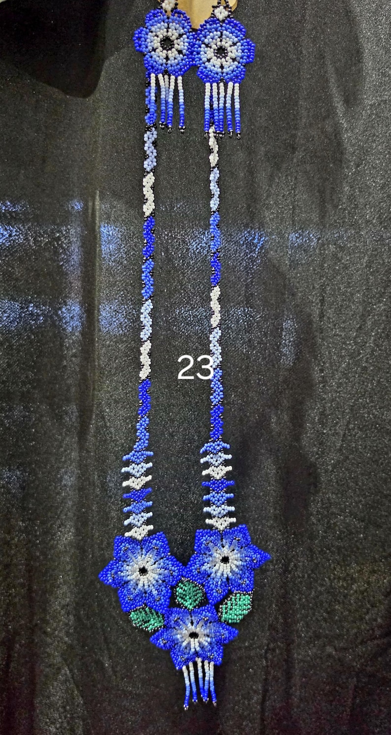 May include: A long beaded necklace featuring a floral pendant with three blue, white, and green flowers. The necklace has two long strands of blue and white beads, with two smaller matching flower accents at the top. The number 23 is visible.