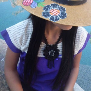 May include: A straw hat with hand-painted blue and orange flowers, paired with a purple and white blouse featuring a textured yoke. A black beaded necklace with a large, circular, flower-shaped pendant and dangling fringe accents rests on the chest.