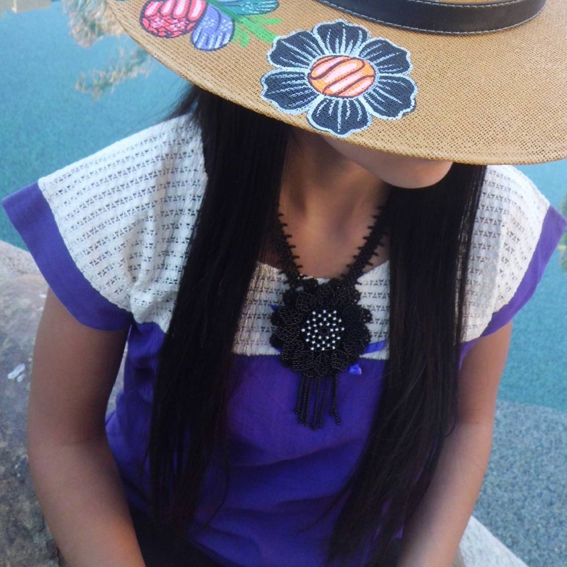 May include: A straw hat with hand-painted blue and orange flowers, paired with a purple and white blouse featuring a textured yoke. A black beaded necklace with a large, circular, flower-shaped pendant and dangling fringe accents rests on the chest.